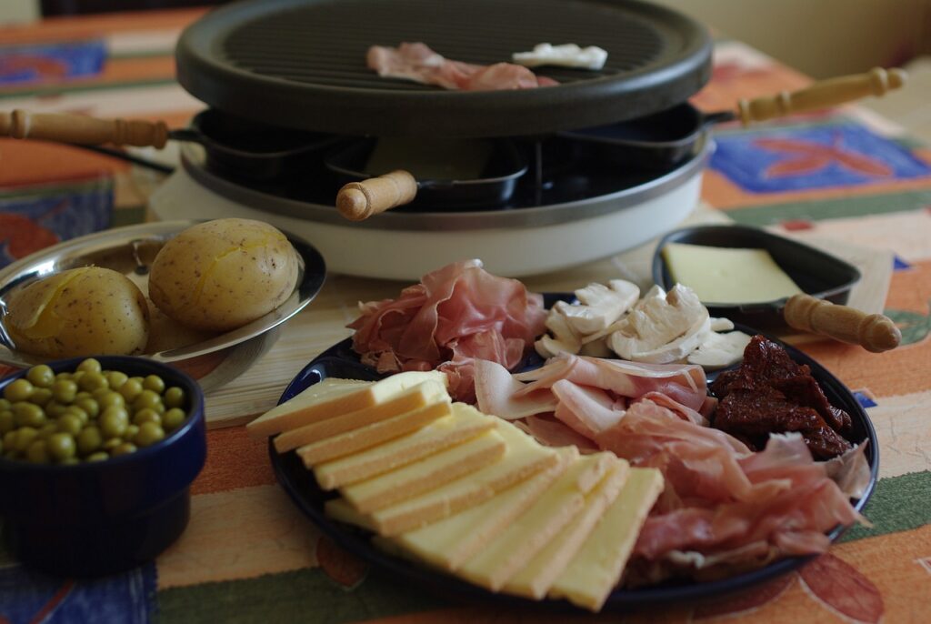 Appareil à raclette dans le fond, avec fromage et plateau de charcuterie et pommes de terres au premier plan